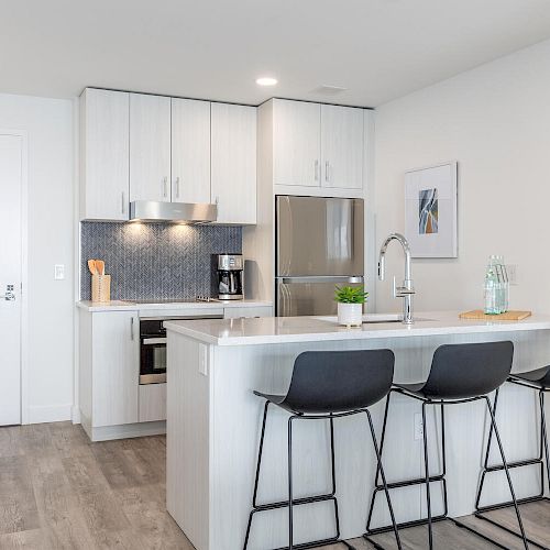 A modern kitchen with a white island, three black stools, stainless steel appliances, and minimalist decor; a framed picture is on the wall.