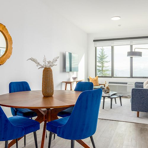 Modern living room with a wooden dining table, blue chairs, a blue sofa, wall-mounted TV, lamp, and a round mirror on the wall.