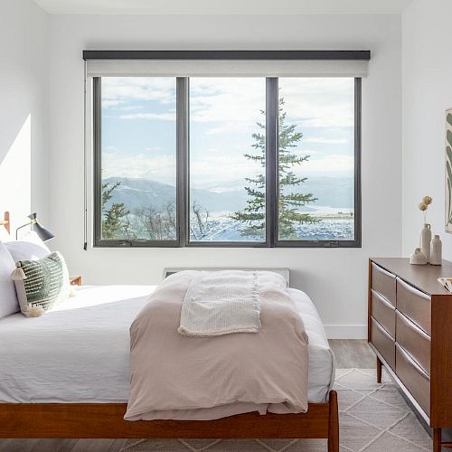 A modern bedroom with a bed, dresser, round mirror, and large window showcasing a scenic view. Minimalist decor and natural light fill the room.