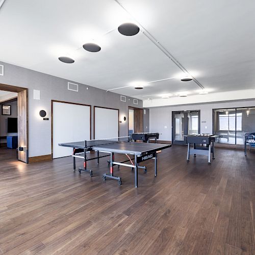 The image shows a spacious game room with ping pong tables, arcade machines, seating areas, and modern lighting fixtures on a wooden floor.