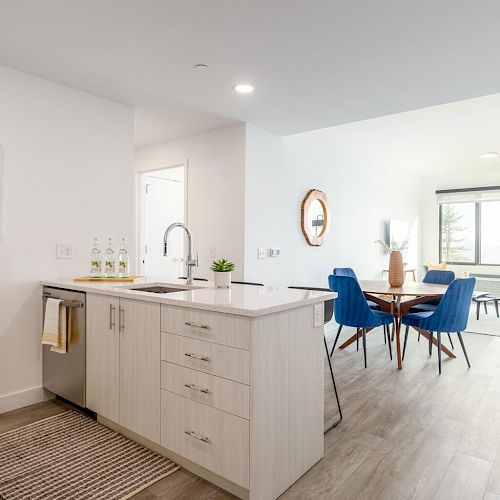 Modern, open-concept kitchen and living area with light wood flooring, a central island, blue dining chairs, and a window letting in natural light.