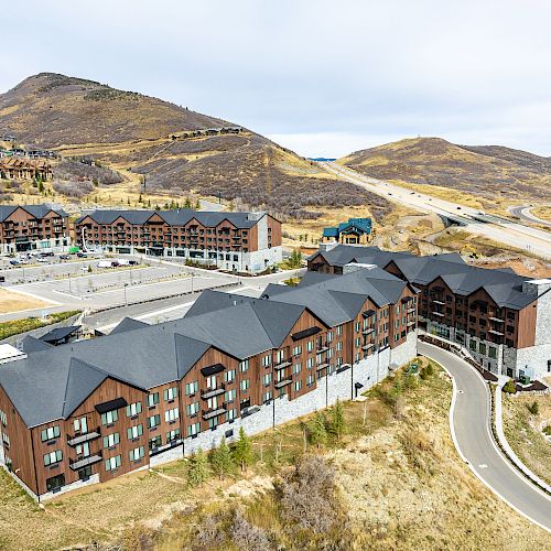 An aerial view of a complex of multi-story buildings with sloping roofs, situated in a mountainous area with winding roads and parking spaces.