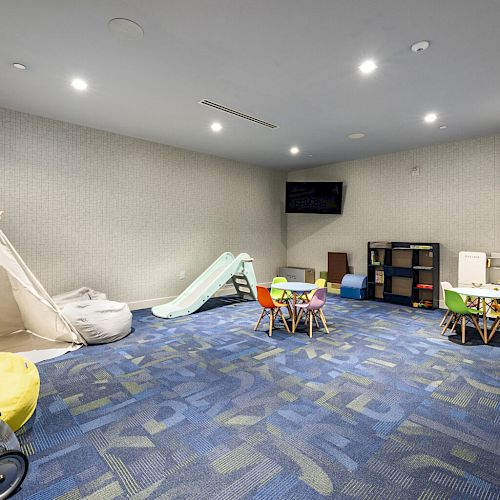 A children's playroom features a tent, slide, chairs, tables, bean bags, shelves, and toys with bright lighting and a blue-patterned carpet.