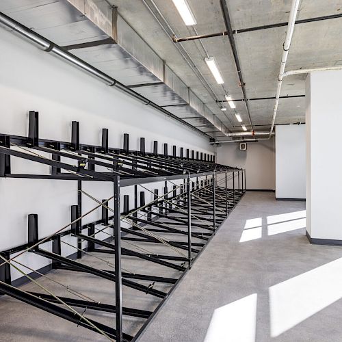This image shows an empty indoor storage area with metal racks, likely for bicycles, along one wall and a large window allowing natural light in.