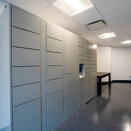 This image shows a narrow corridor with gray lockers on the left side and a small table further down the hall. Fluorescent lights are overhead.