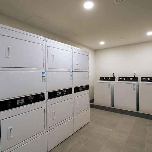 The image shows a laundry room with several stacked washing and drying machines on the left, and three single washing machines on the right.