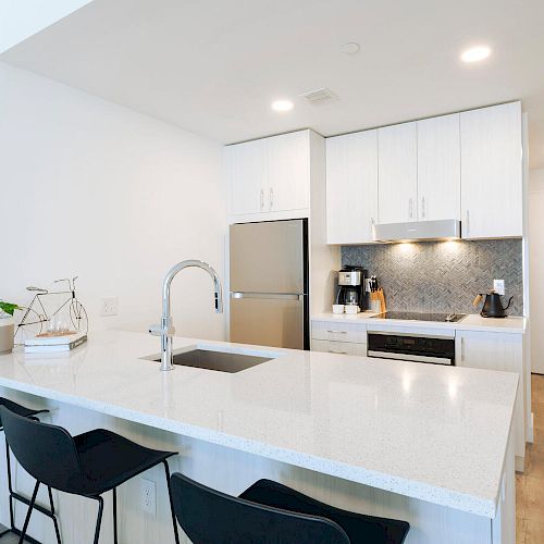 A modern kitchen with white cabinets, stainless steel appliances, a kitchen island with a sink, three black barstools, and various decor items.