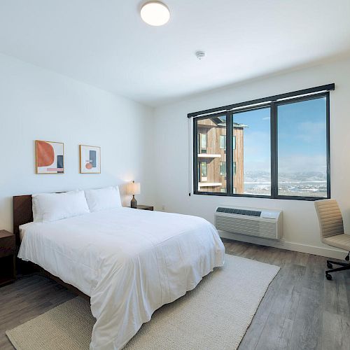 A modern bedroom featuring a bed with white linens, two bedside tables with lamps, a desk with a chair, and a large window with a city view.