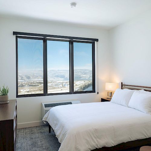 A modern bedroom features a bed with white linens, a window with a scenic view, a dresser, a TV, and a small nightstand with a lamp.