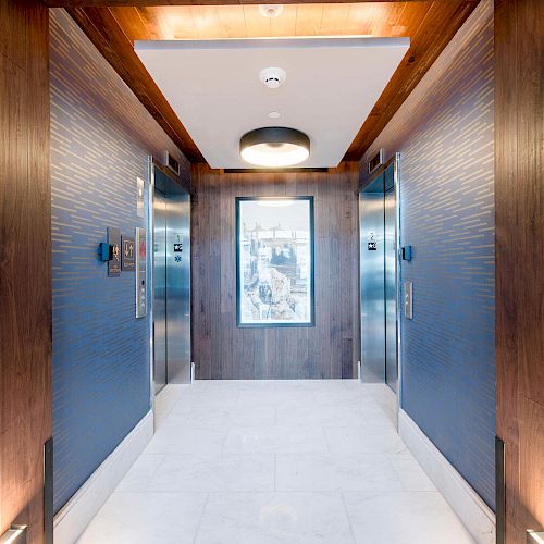 The image depicts a modern elevator lobby with two elevators, blue walls, wooden paneling, and a central overhead light fixture, ending the sentence.