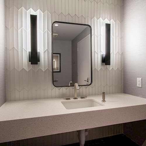 A modern bathroom vanity features a rectangular mirror, geometric wall tiles, two vertical lights, a sink, and a minimalist countertop.