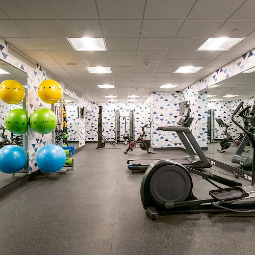 This image shows a gym with exercise equipment such as an elliptical machine, stability balls, and strength training machines, with mirrored walls.