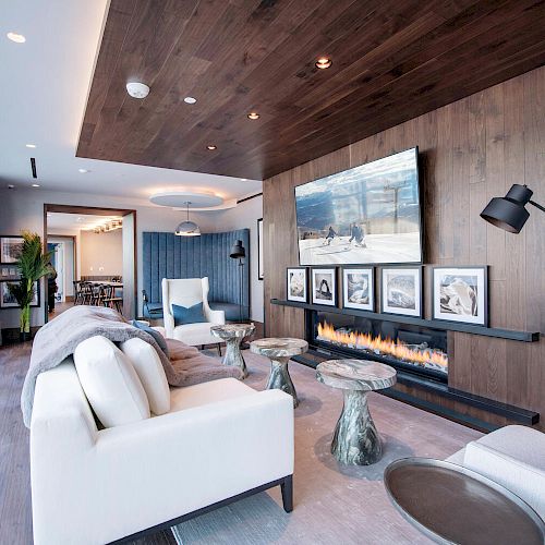 A modern living room with white furniture, a fireplace, wall-mounted TV, large windows, and framed photos.
