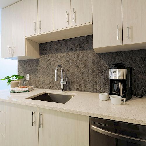 A modern kitchen with light wood cabinets, black backsplash, stainless steel sink, coffee maker, and two cups on a white countertop.