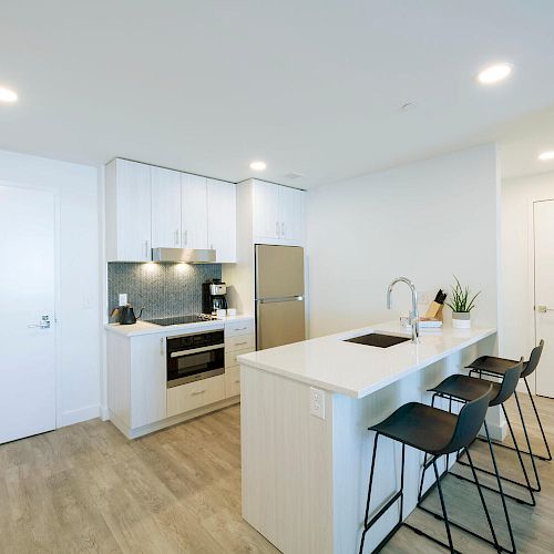 A modern kitchen with light wood floors, white cabinets, a stainless steel refrigerator, a stove, and a large island with three black barstools.