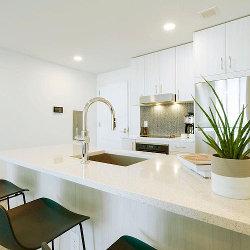 This image shows a modern kitchen with a large island, two black chairs, a plant, and a knife set on the counter, and white cabinetry.