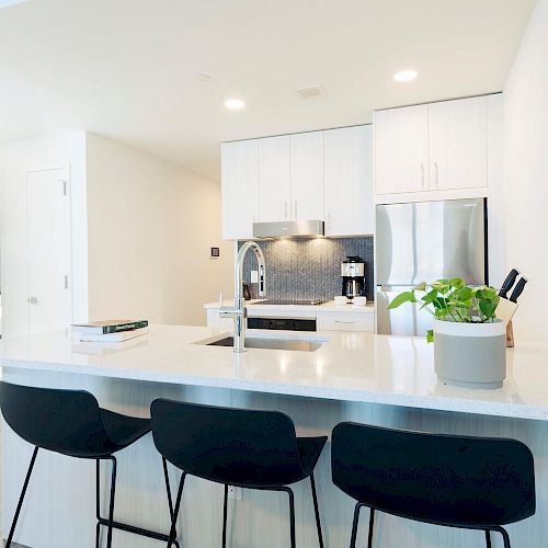 A modern kitchen with white cabinetry, a stainless steel fridge, and a counter with three black stools and a plant. Ending the sentence.
