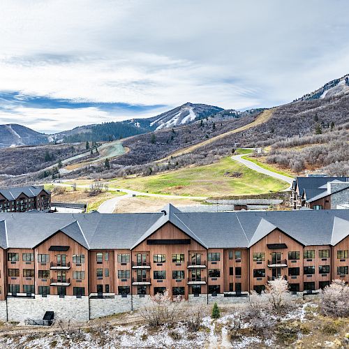 A large building complex is situated in a mountainous area with ski slopes and hills in the background under a partly cloudy sky.