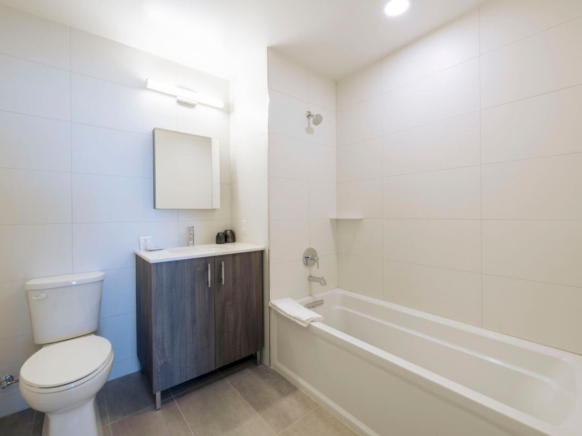 A modern bathroom with a toilet, vanity with a mirror, and a bathtub with a showerhead, featuring a minimalistic design and neutral colors.