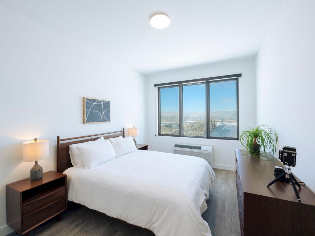 A bright bedroom with a large bed, nightstands with lamps, a dresser with a plant and decor, and a window with a city view.