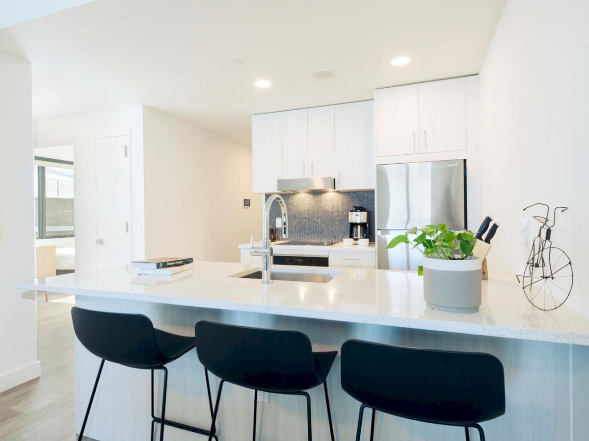 A modern kitchen with a white island, three black stools, stainless steel appliances, and a plant on the counter. End of description.