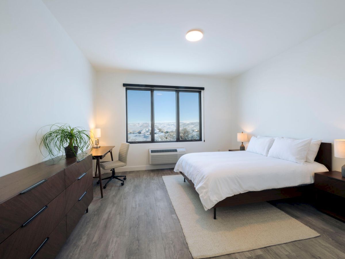 A modern bedroom with a double bed, two bedside tables with lamps, a large dresser with a plant, a desk, and a window with a city view.