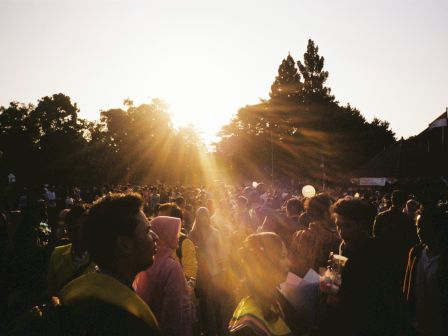A crowd of people stands outdoors at sunset, with sunlight filtering through trees, giving the scene a warm, golden glow.