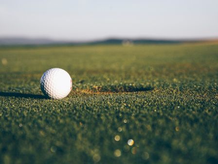 A close-up view of a golf ball sitting near a hole on a golf course, with the fairway and horizon blurred in the background.