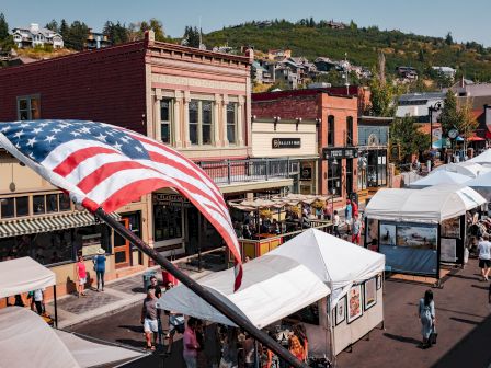 A bustling street fair in a small town features vendor tents, an American flag, and historic brick buildings with a scenic hill in the background.