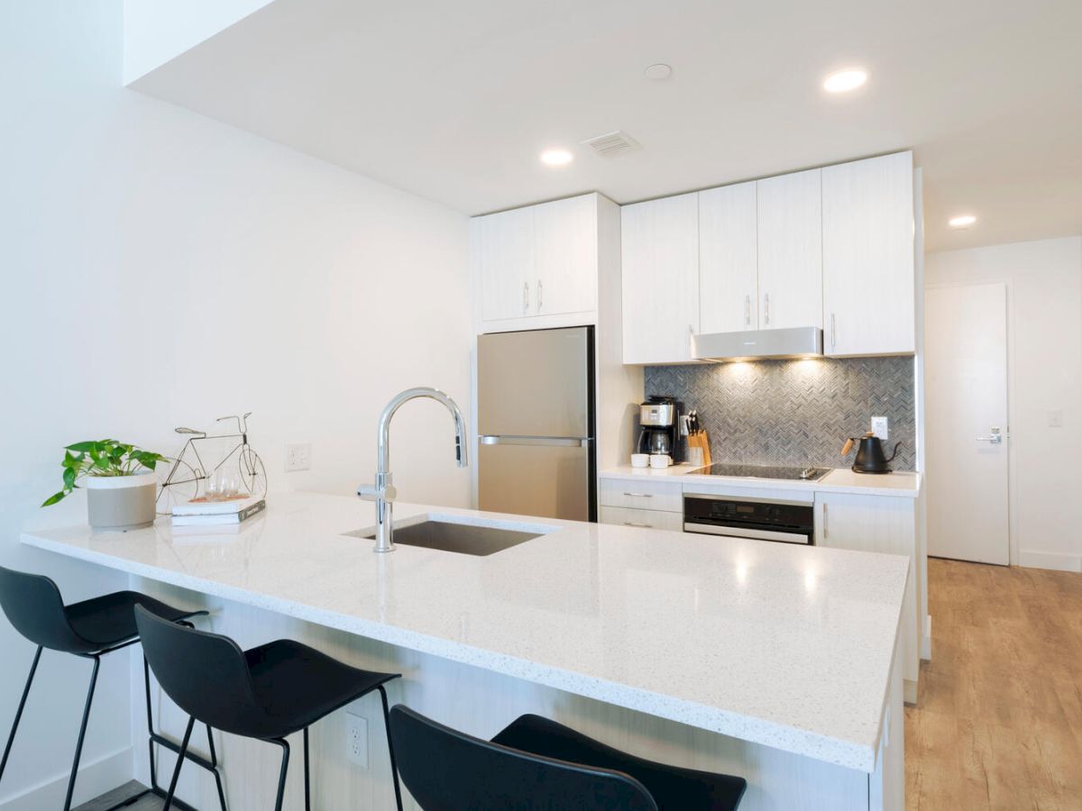 A modern kitchen with a white countertop, black chairs, stainless steel appliances, and minimalist decor items, including a small plant, ending the sentence.