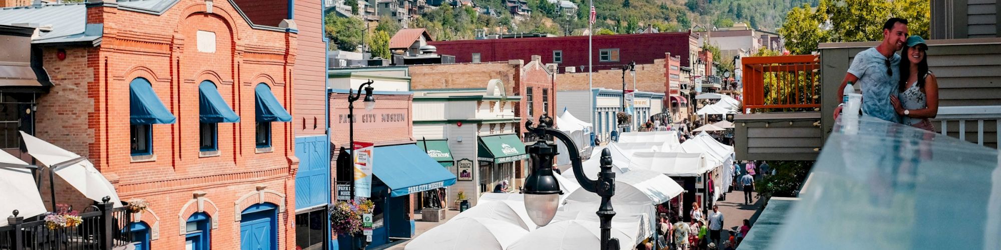 A bustling street fair in a scenic town with colorful buildings, tents, and people enjoying the event on a sunny day in a mountainous area.