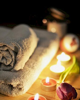 The image shows rolled towels, lit candles, and a purple tulip, creating a relaxing spa-like atmosphere on a wooden surface.