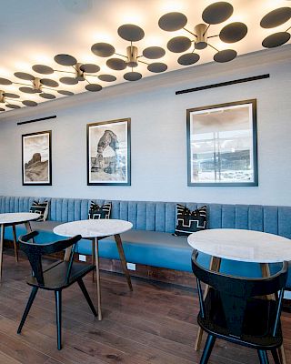 A modern seating area with round tables, black chairs, blue bench seating, decorative pillows, framed artwork on the wall, and unique ceiling lights.