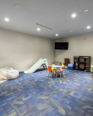 A children's playroom with a small tent, play slide, tables, chairs, toy shelves, and a TV on the wall. The floor is carpeted in blue.
