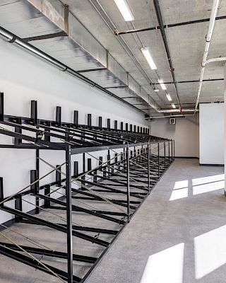 This image shows an empty industrial-style room with multiple rows of black metal racks, likely for storage, and large windows on one side.