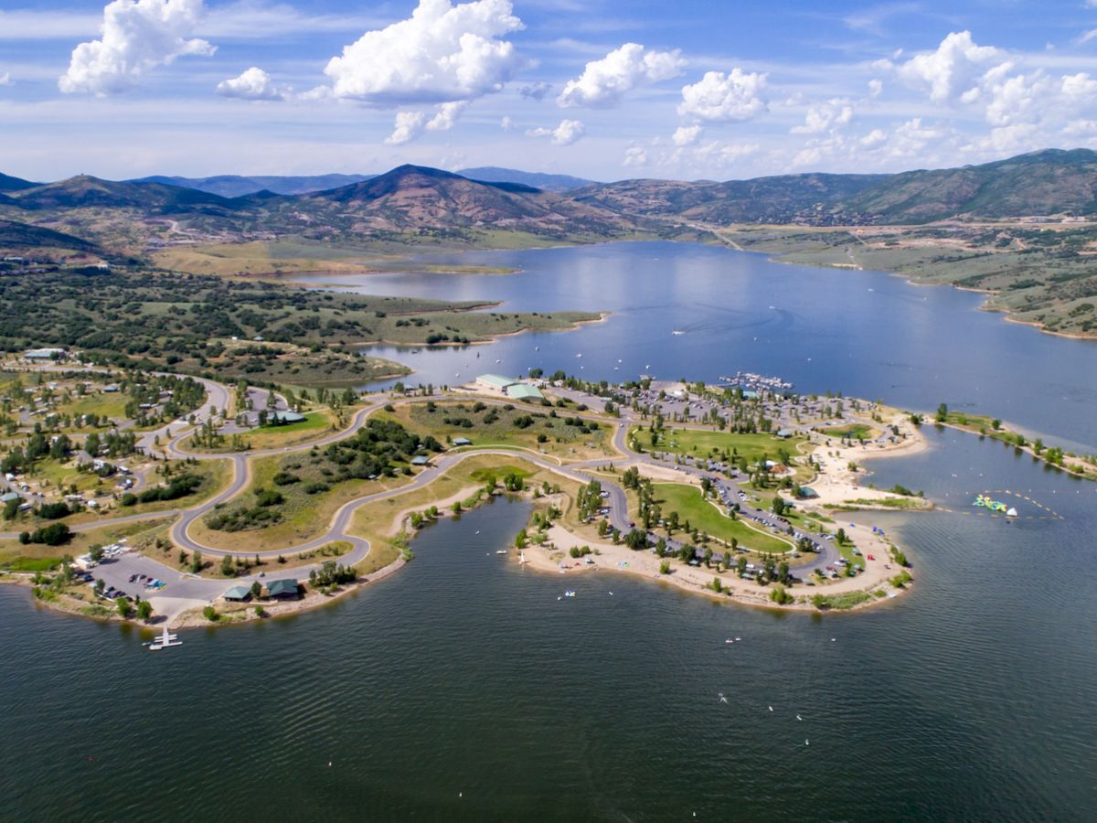 Aerial view of a scenic lake surrounded by a hilly landscape with winding roads, campgrounds, and scattered vegetation, all under a clear, partly cloudy sky.