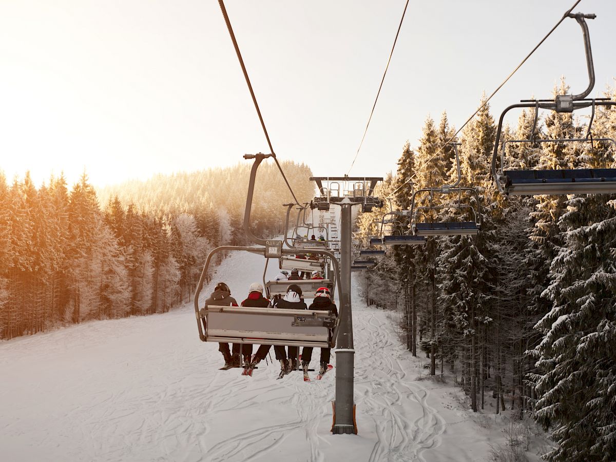 Skiers on a chairlift ascend a snow-covered mountain surrounded by snow-laden trees during a serene sunset.