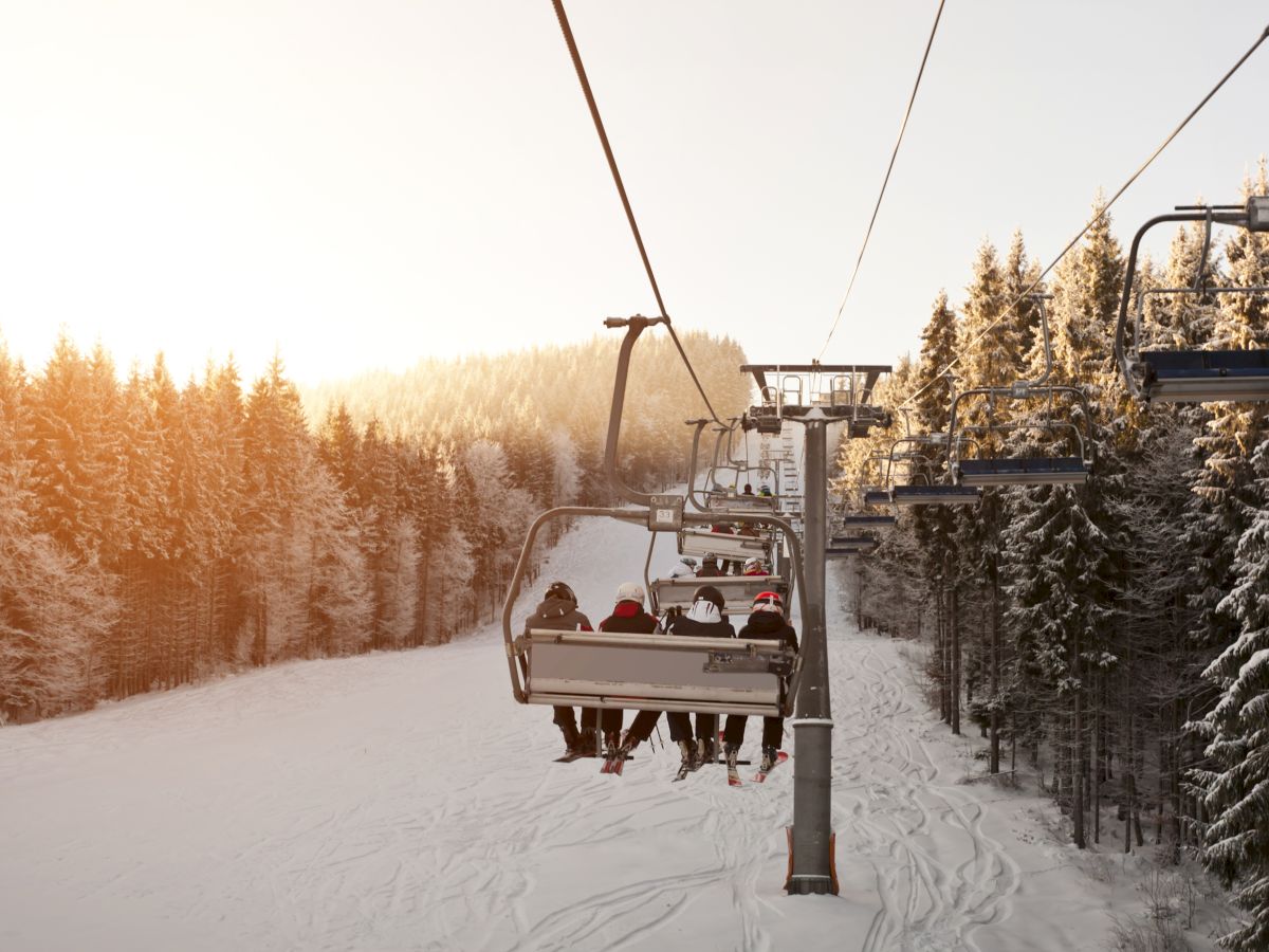 A ski lift carrying people over a snowy landscape with forested slopes and a sunset in the background.